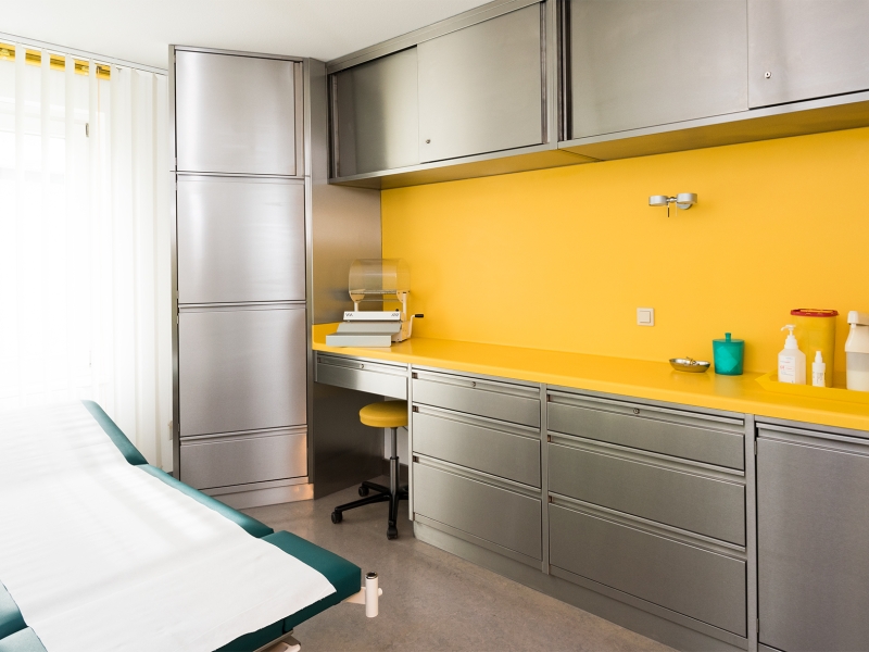Ordinationsraum mit Edelstahl-Wandverbau (links: Hochschrank, oben: 2x Wandhängeschrank mit Schiebetür, unten: Arbeitsschränke teilweise unterfahrbar für Hocker) mit Corian-Abdeckung